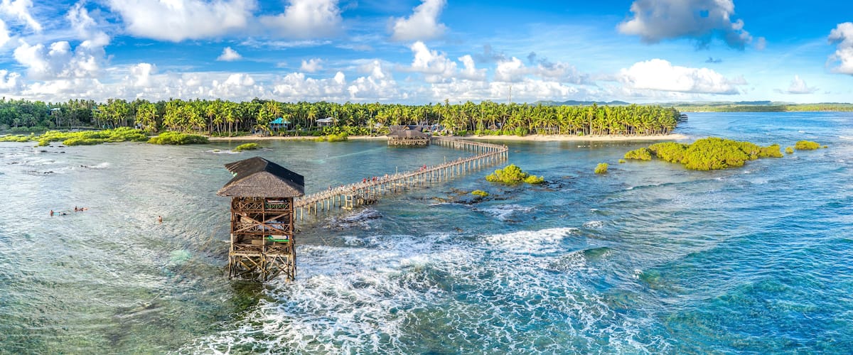 A tropical island with a wooden pier leading to a thatched-roof structure, surrounded by palm trees and clear blue waters.