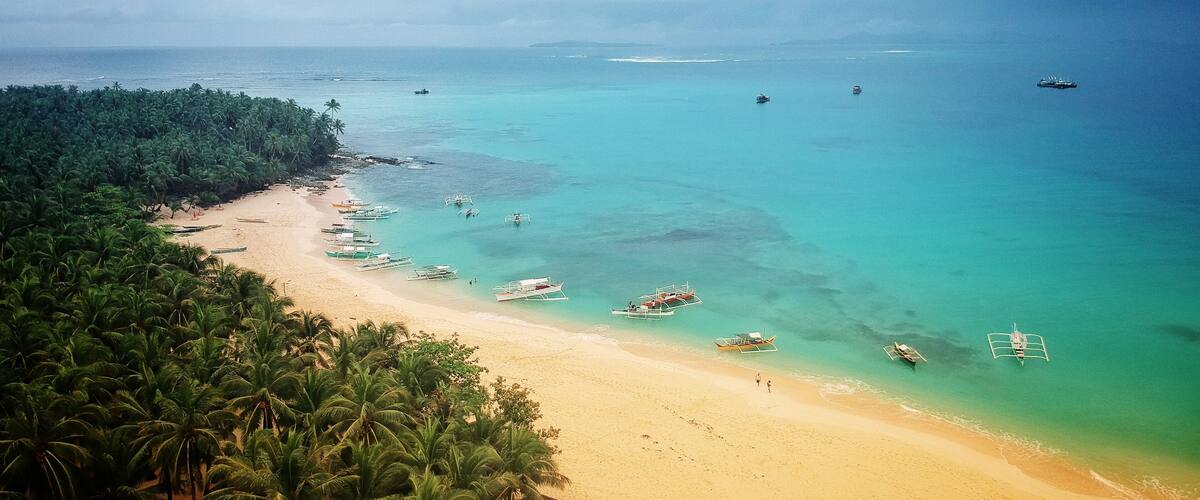 A tropical beach with clear turquoise waters, palm trees, and anchored boats.