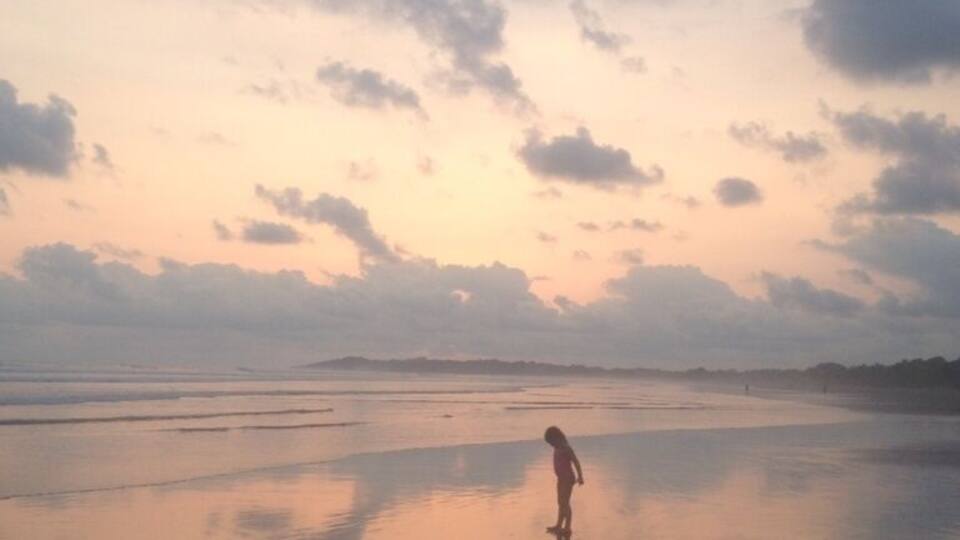 A person standing on a wet beach at sunset with a cloudy sky.