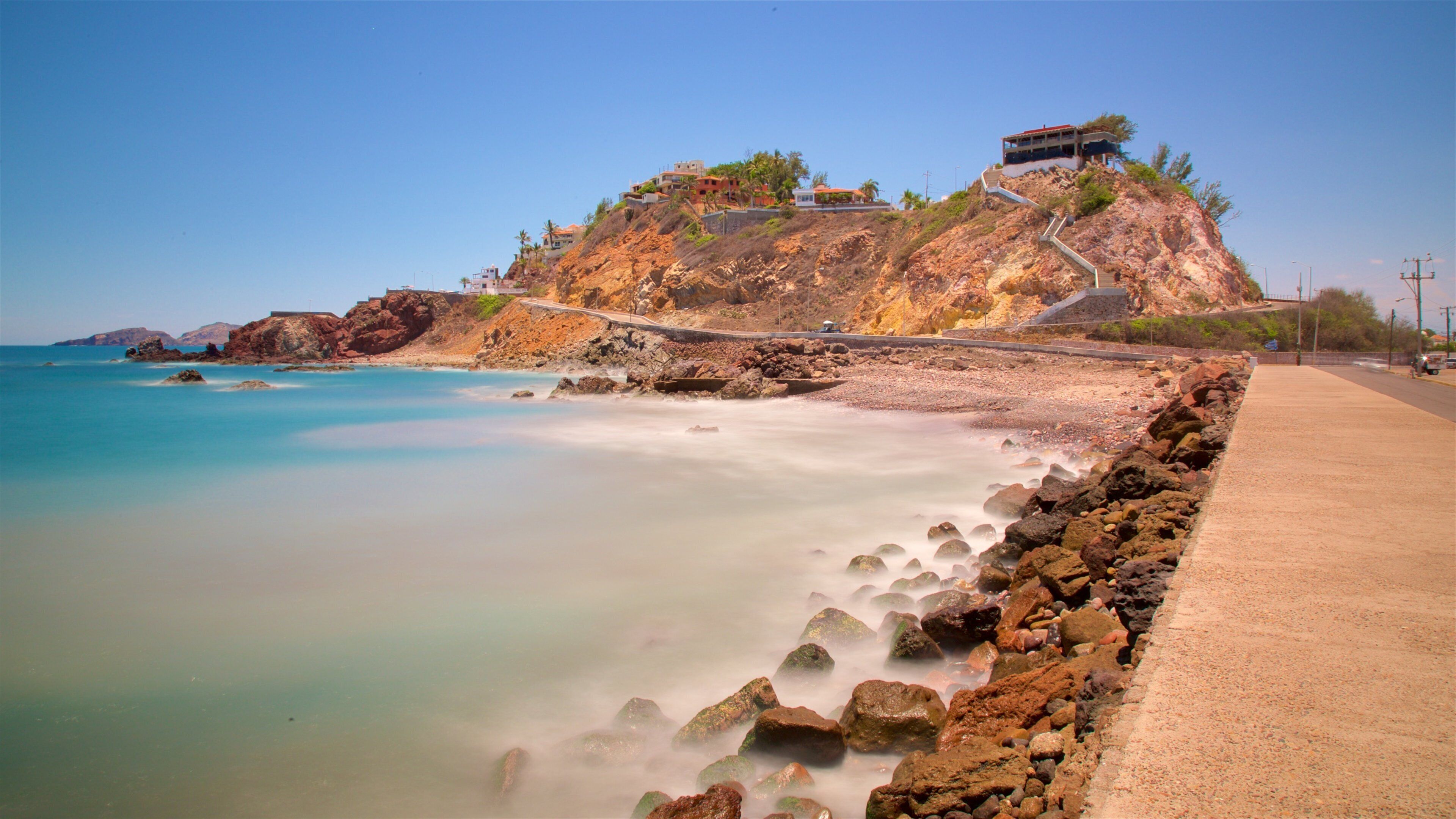 A coastal landscape with a rocky beach, a clear blue sea, and a hillside with buildings.