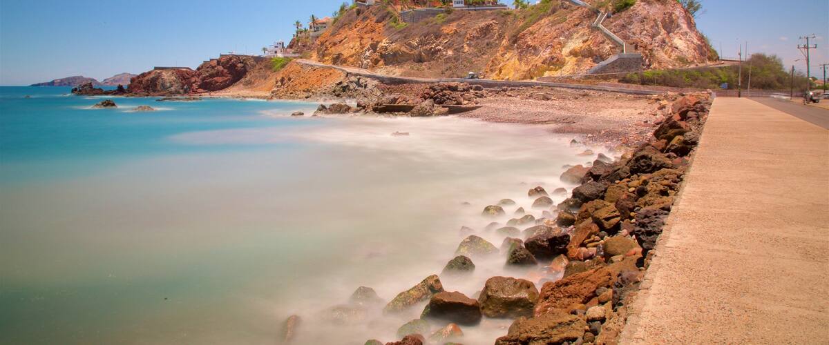 A coastal landscape with a rocky beach, a clear blue sea, and a hillside with buildings.