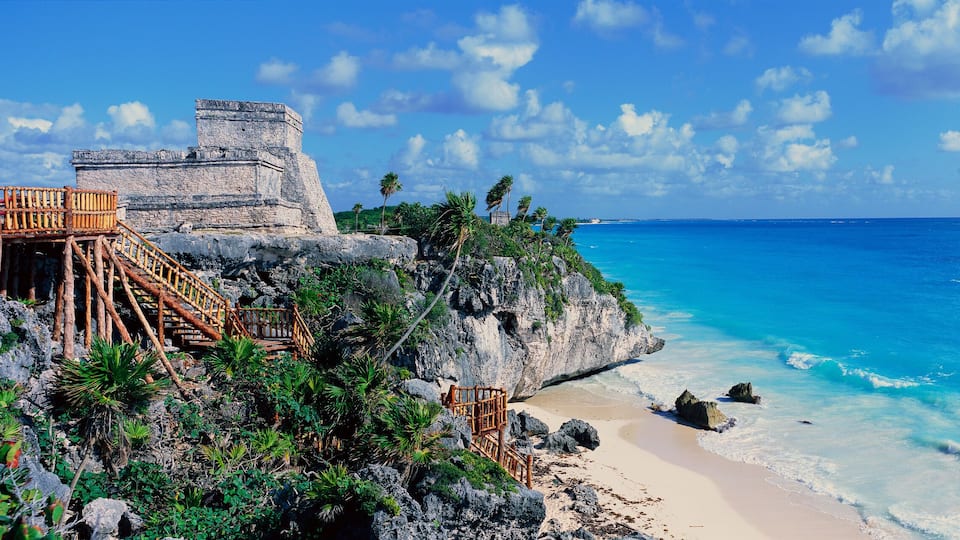 A coastal scene with a stone structure, wooden staircase, beach, and clear blue ocean.