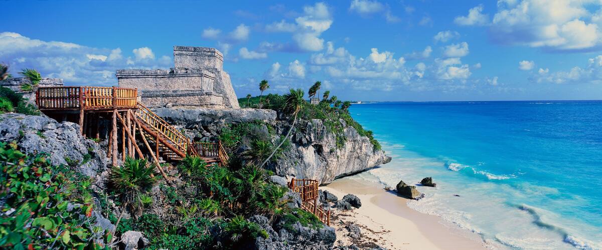 A coastal scene with a stone structure, wooden staircase, beach, and clear blue ocean.