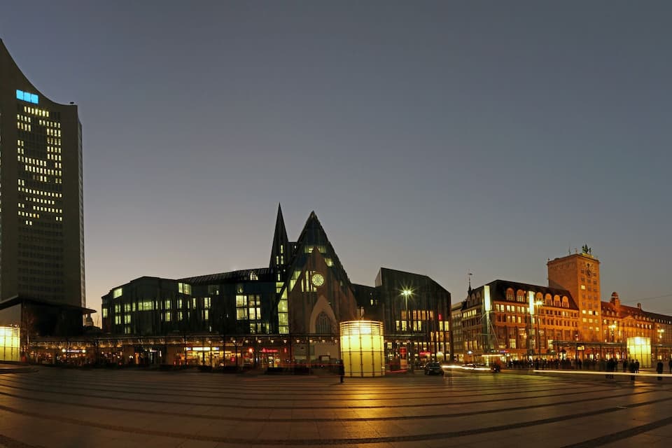 A city square with modern buildings, a church, and a clock tower at dusk.