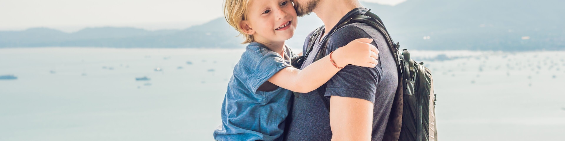 A person holding a child by a waterfront with mountains in the background.