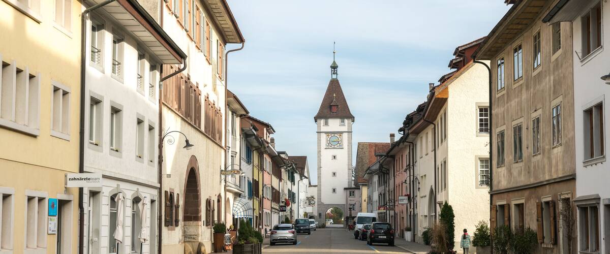 A street with buildings, a clock tower, and parked cars.