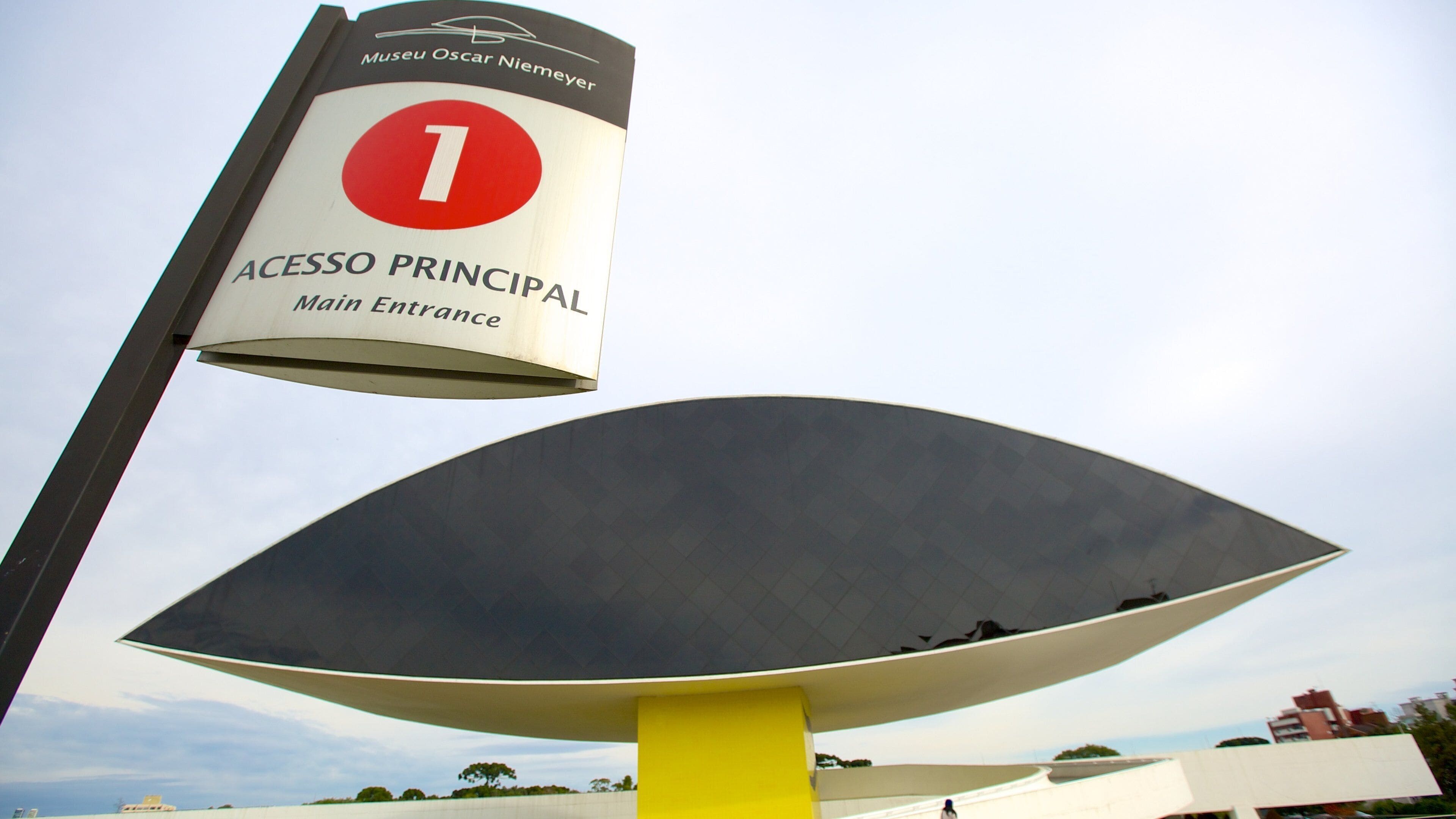 A large, modern sculpture with a yellow base and a red and white sign above it reading "Museu Oscar Niemeyer" and "Acesso Principal Main Entrance".