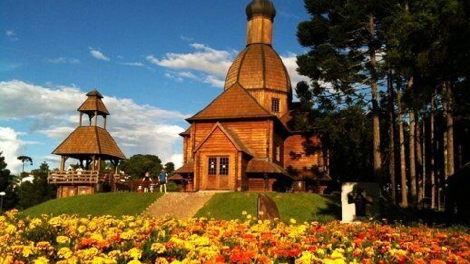 A wooden church with a bell tower and a gazebo surrounded by colorful flowers.
