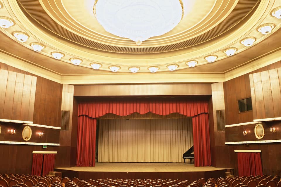 Un auditori amb un escenari, cortines vermelles, una ostentosa pèndol i files de seients.