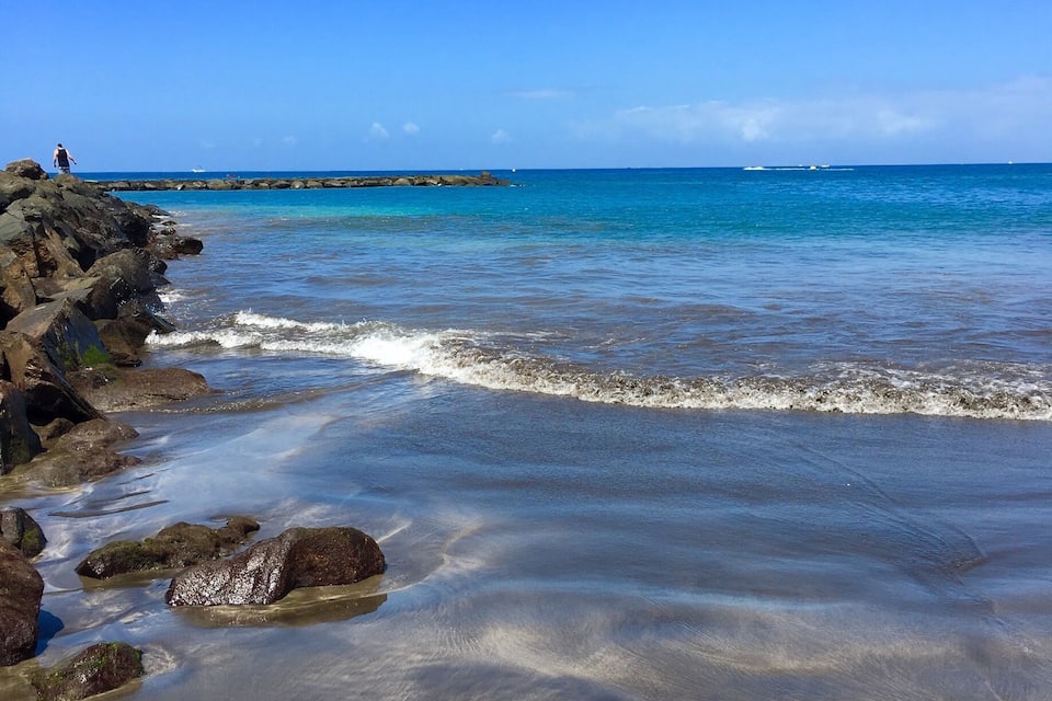 Una playa con agua azul cristalina, costa rocosa y un muelle a lo lejos.