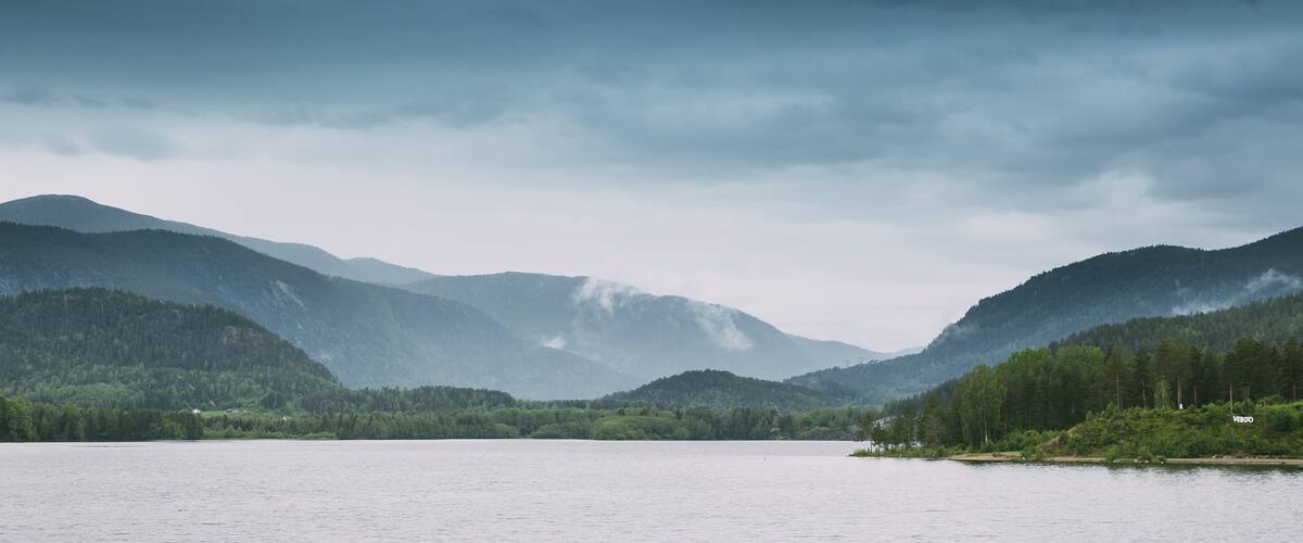 A calm lake surrounded by forested mountains under a cloudy sky.