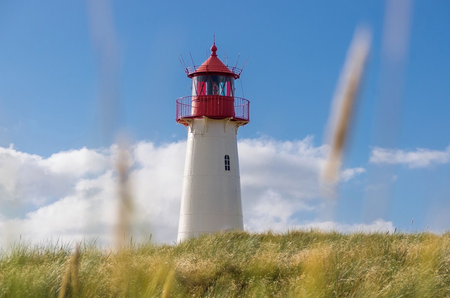 Un faro con la parte superior roja y la base blanca, rodeado de hierba y nubes.