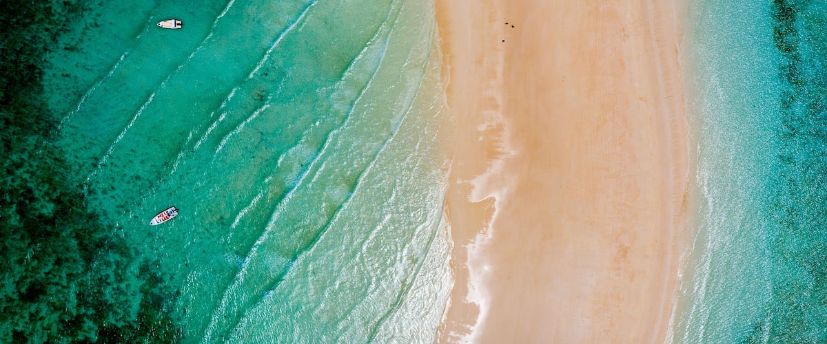 An aerial view of a beach with clear turquoise waters, a sandy shore, and a few boats.