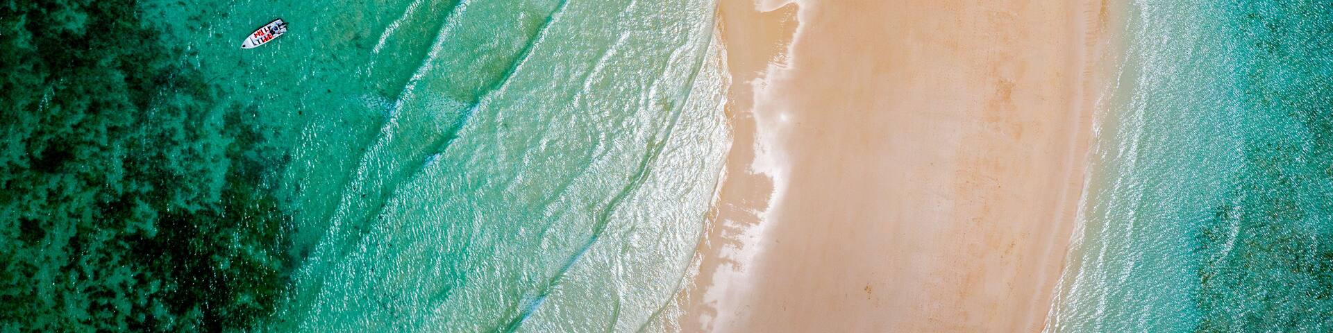 An aerial view of a beach with clear turquoise waters, a sandy shore, and a few boats.