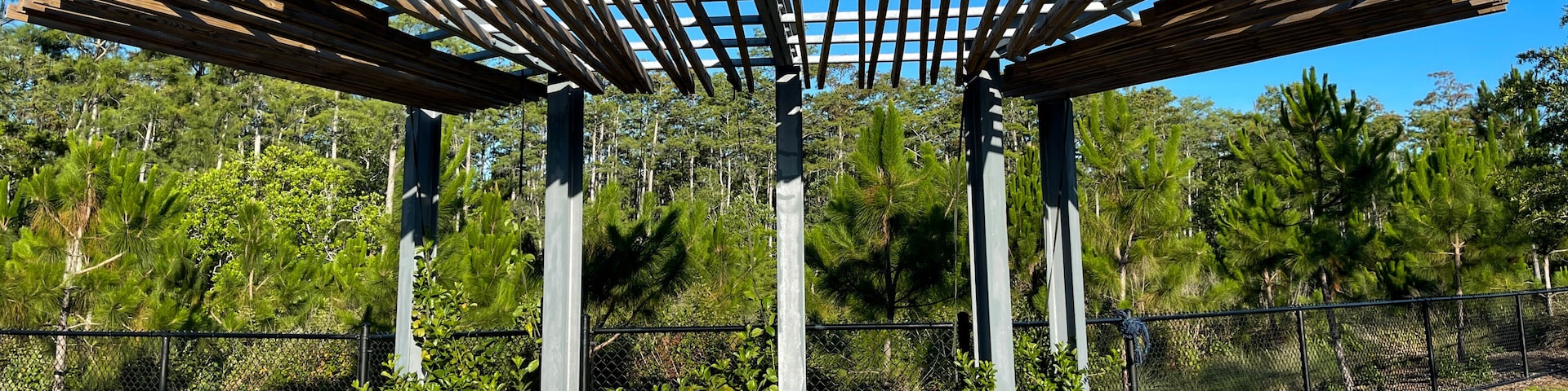 A wooden pergola structure with a concrete base, surrounded by a chain-link fence and lush greenery.