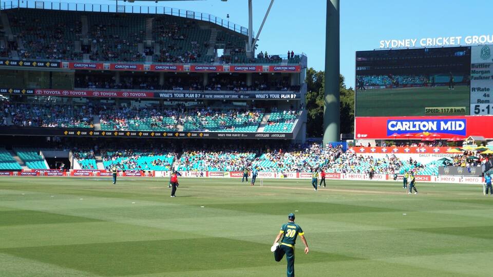 A cricket stadium with multiple tiers of seating, advertisements, and a large screen displaying a game.