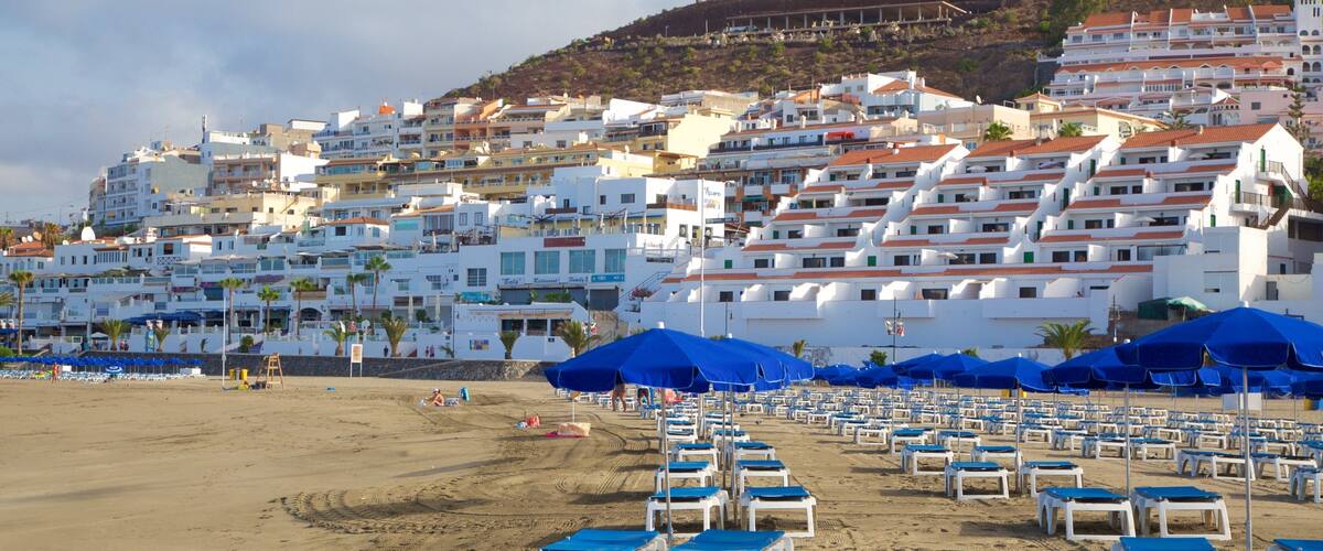 A beach with rows of blue and white sun loungers and umbrellas, a sandy shore, and a backdrop of white buildings with red roofs on a hillside.