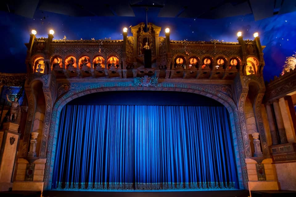 Um teatro com arquitetura elaborada, uma grande cortina azul e iluminação decorativa.