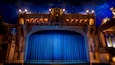 Un théâtre à l’architecture richement décorée, doté d’un grand rideau bleu et d’un éclairage ornemental.