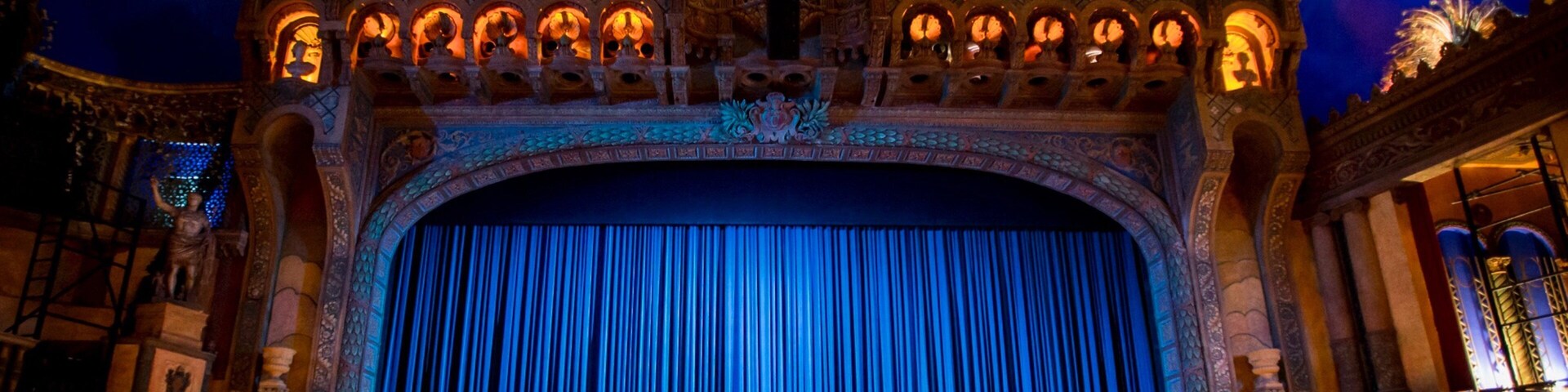 Un teatro con arquitectura ornamentada, un amplio telón azul y una iluminación decorativa.