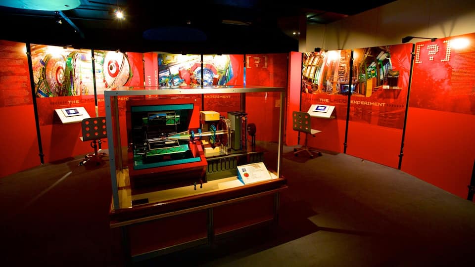 An exhibition with a central display case containing scientific equipment, surrounded by informational panels and a red-lit environment.