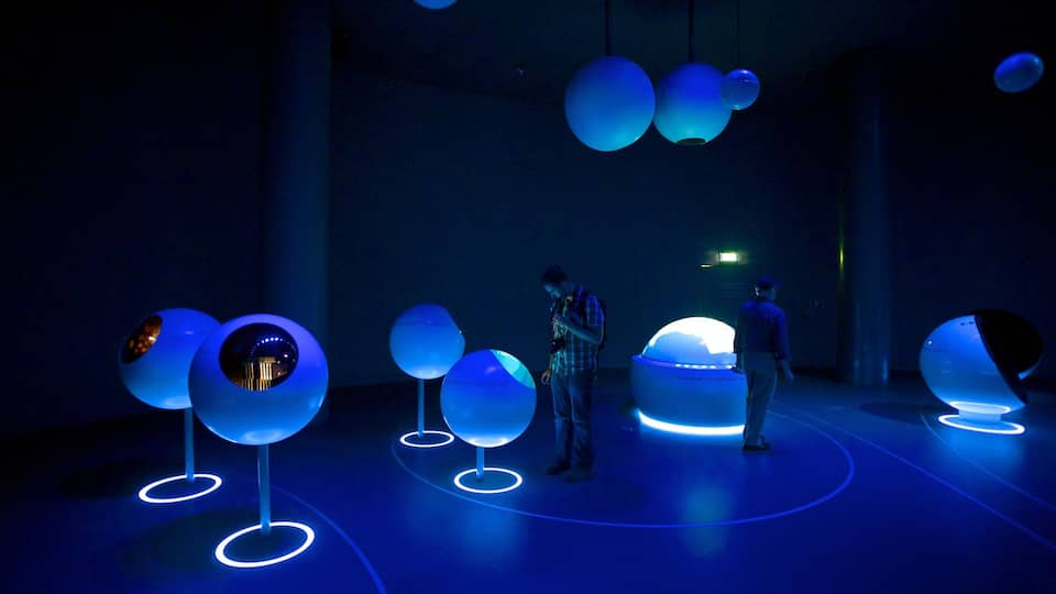 A dark room with illuminated spherical objects and a person standing in front of a large, glowing sphere.
