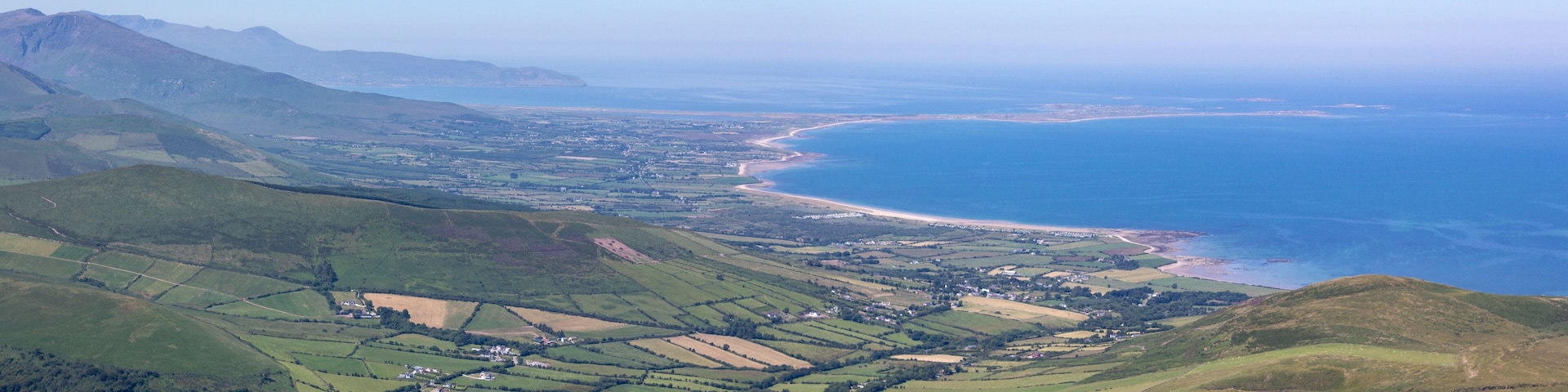 Un paisaje costero con colinas ondulantes, una playa y una cadena montañosa distante.