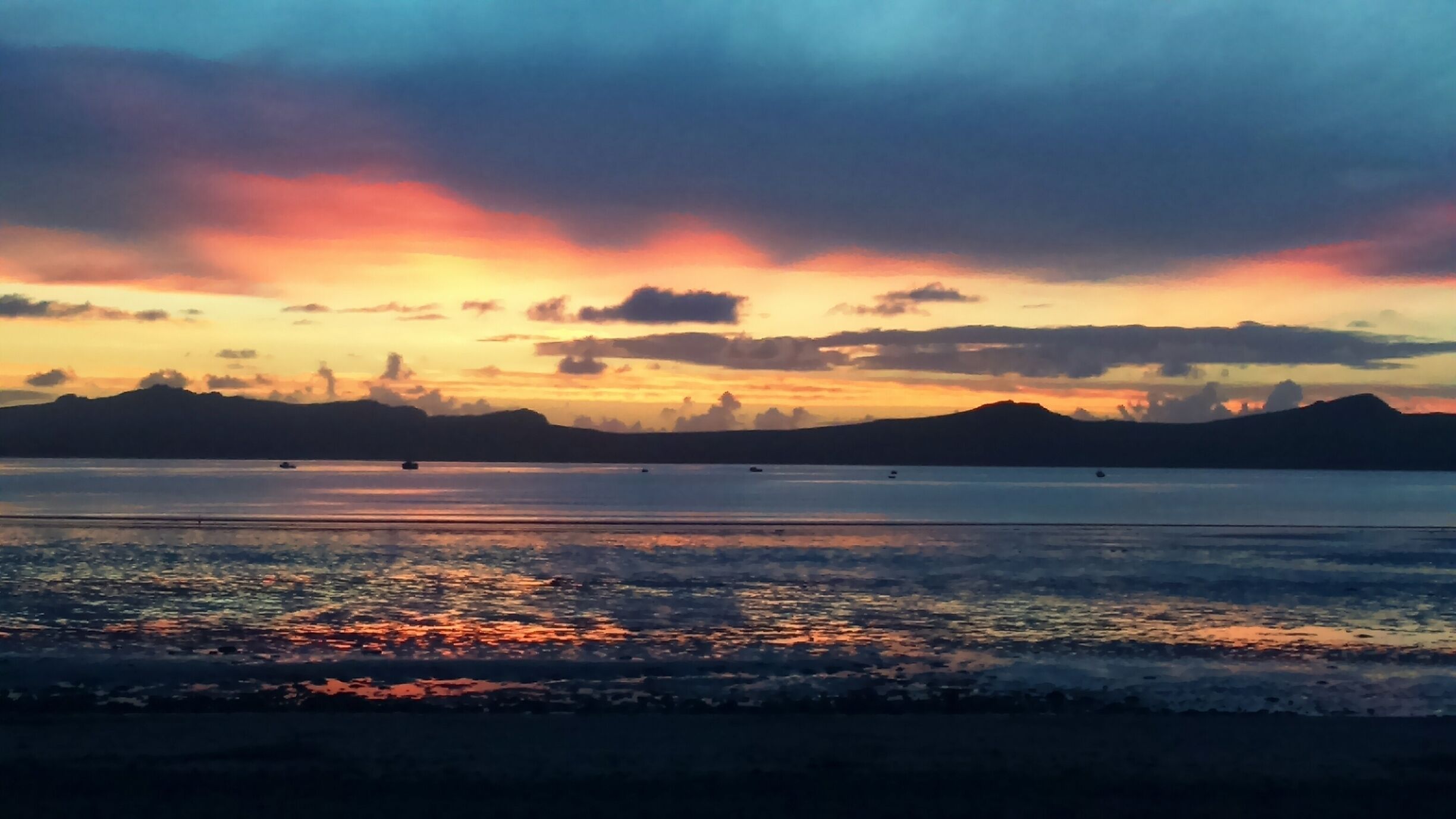 A sunset over a calm body of water with boats and distant mountains.