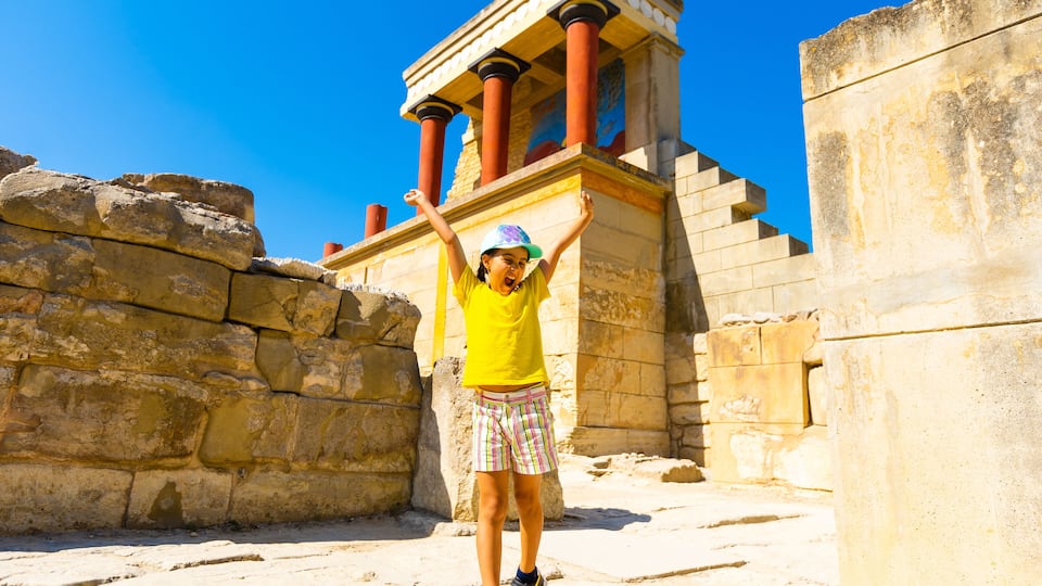A person in a yellow shirt and plaid shorts standing in front of ancient ruins with columns and a staircase.