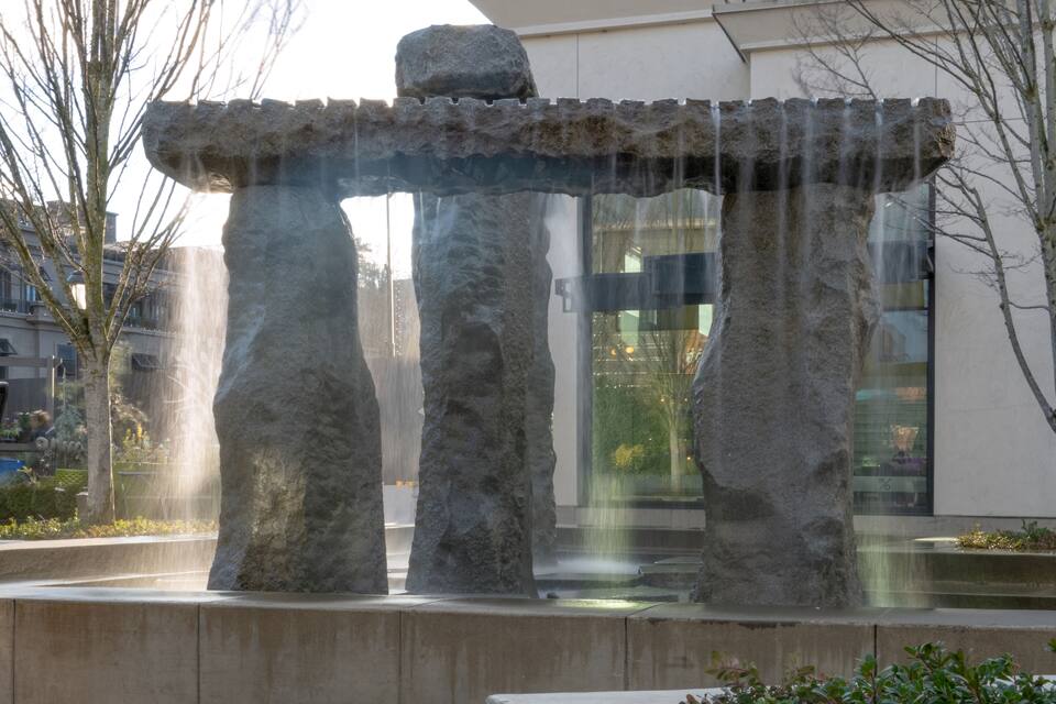 A stone structure with water flowing through it, set in an urban environment with buildings in the background.