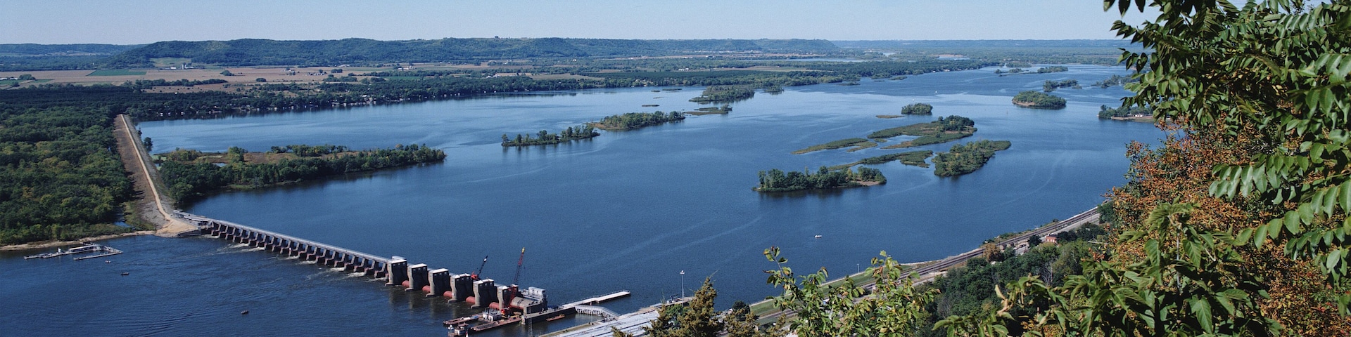 A river with a dam, surrounded by greenery and a clear sky.