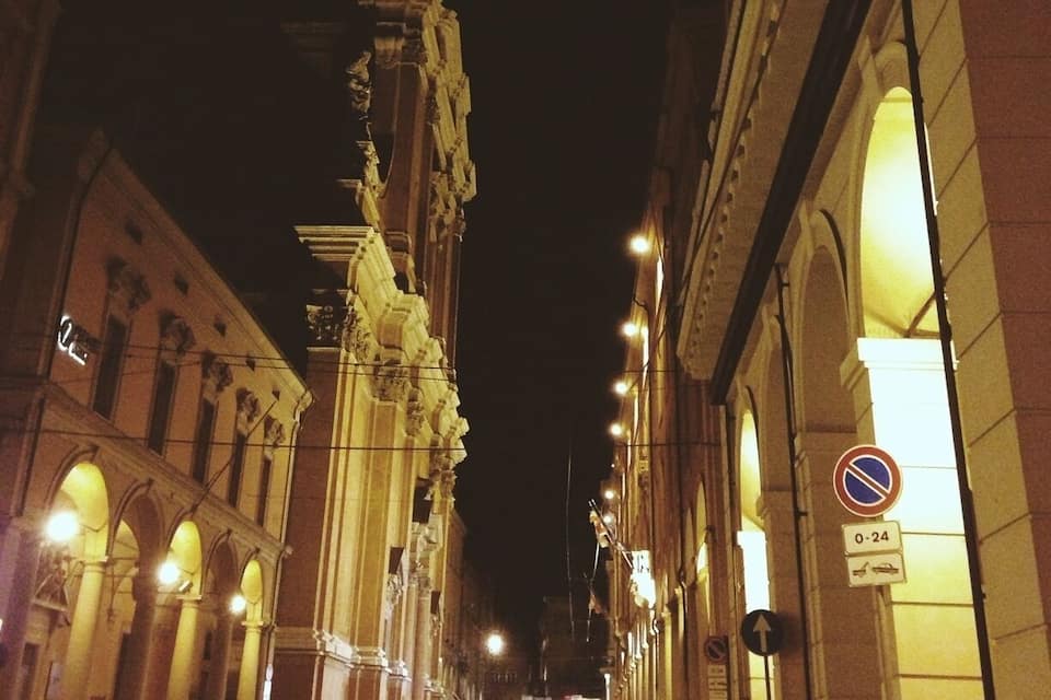 Cena noturna de rua com arquitetura clássica, incluindo uma estrutura imponente com colunas e edifícios iluminados.