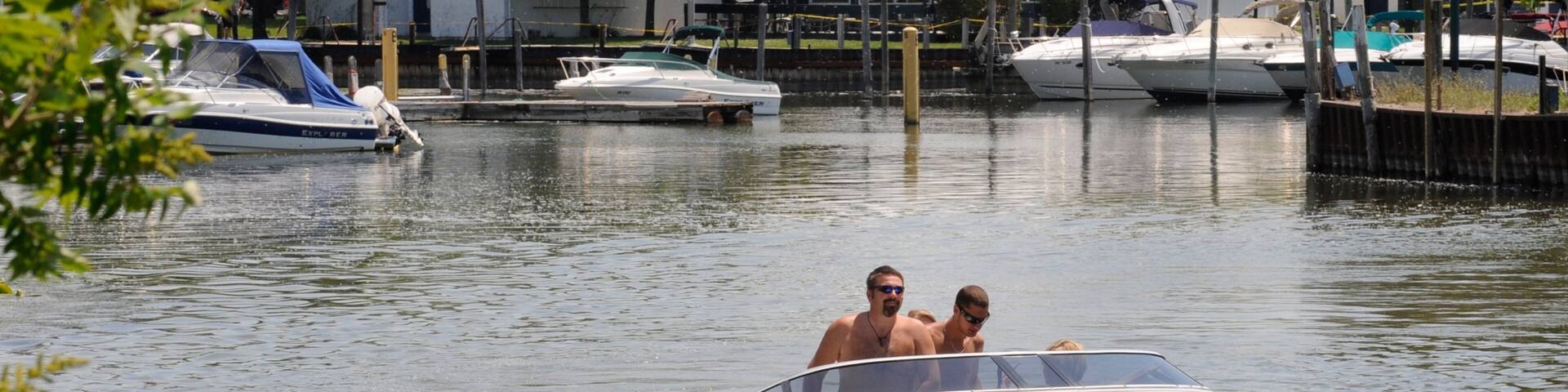 A speedboat with a checkered pattern and the text "BASE THIS PACH" on its side, moving through a marina with other boats docked in the background.