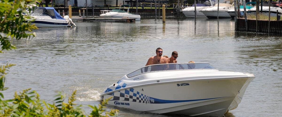 A speedboat with a checkered pattern and the text "BASE THIS PACH" on its side, moving through a marina with other boats docked in the background.