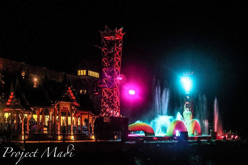 A night scene with illuminated structures, including a tower and domed buildings, and a water fountain display.