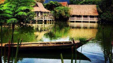 A calm lake with thatched-roof huts, lush greenery, and a wooden boat.