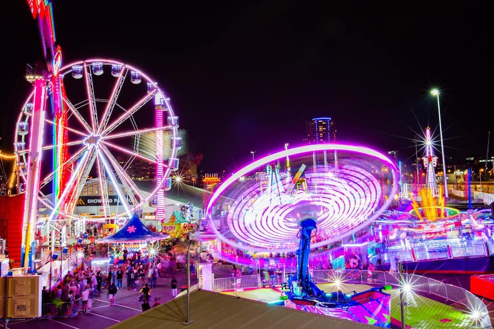 Un parque de atracciones iluminado con intensidad en la noche, con dos grandes norias y diversas atracciones.