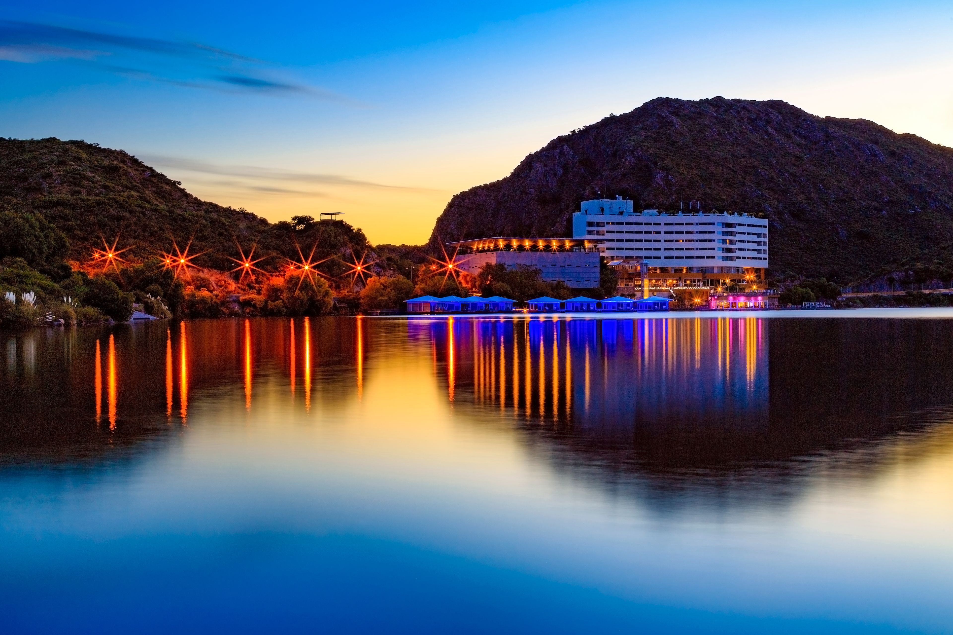 Una orilla con un edificio reflejado en aguas tranquilas, rodeada de colinas.