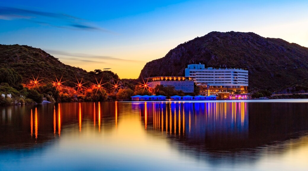 A waterfront with a building reflecting on calm water, surrounded by hills.