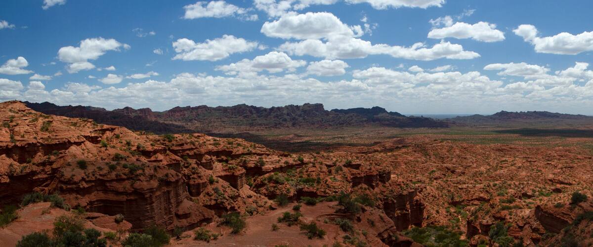 A vast desert landscape with red rock formations and sparse vegetation under a partly cloudy sky.