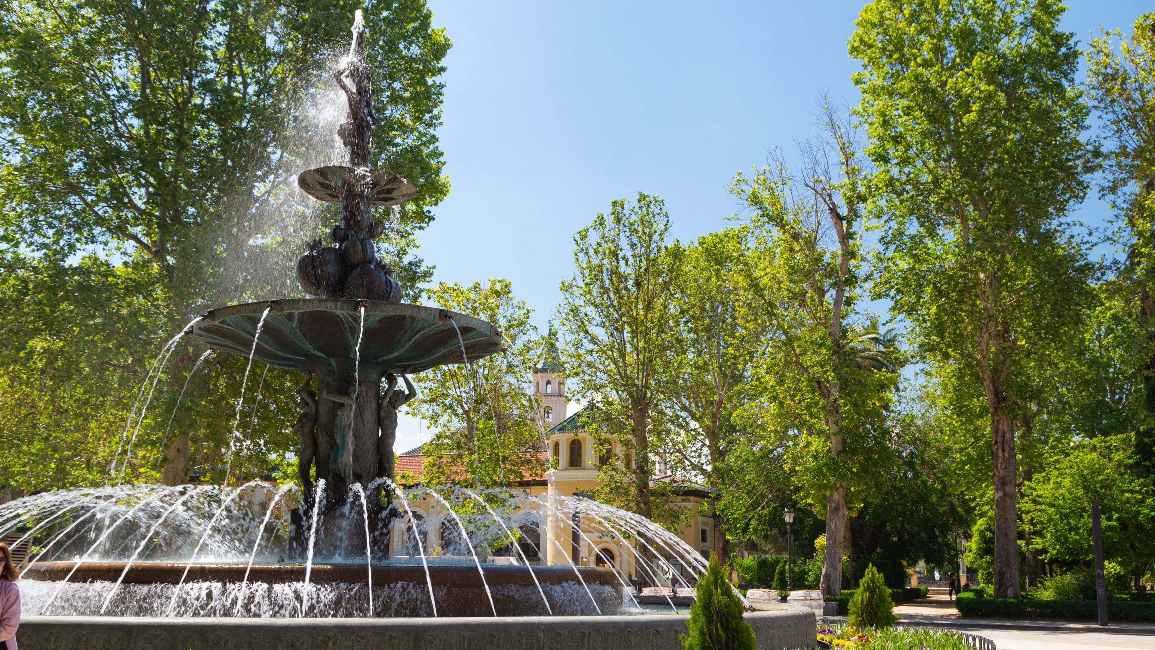 A fountain with multiple water jets, surrounded by lush green trees and a building with a tower in the background.