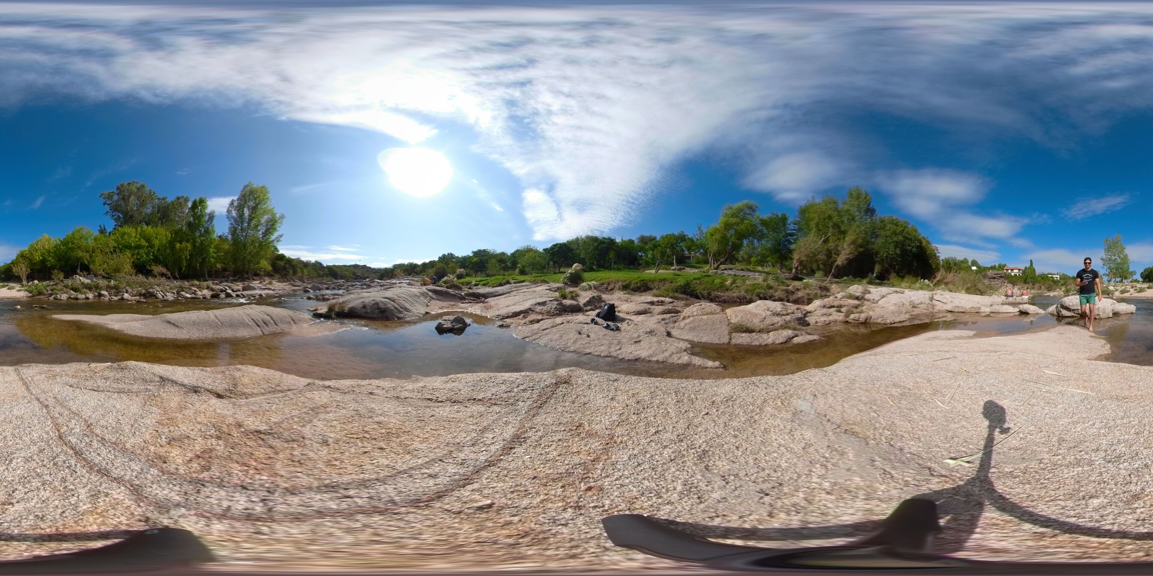 A panoramic view of a rocky riverbed with clear water, surrounded by greenery and trees.
