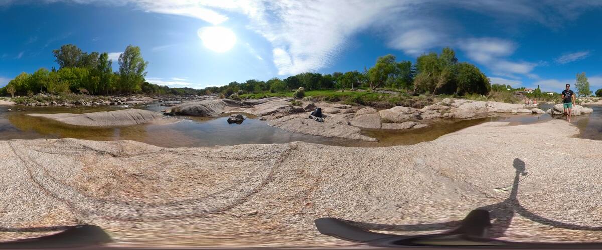 A panoramic view of a rocky riverbed with clear water, surrounded by greenery and trees.