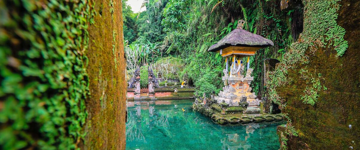 A serene water body with a traditional thatched-roof structure, surrounded by lush greenery and stone architecture.