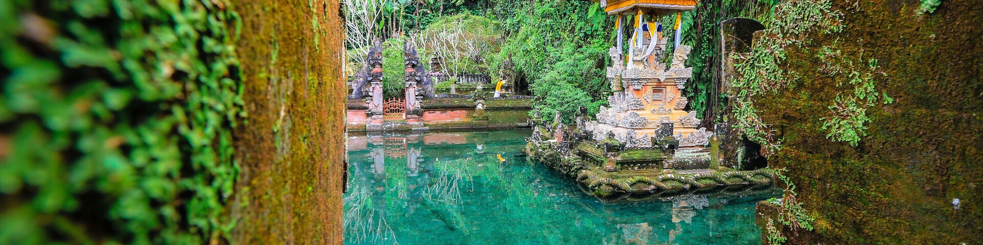 A serene water body with a traditional thatched-roof structure, surrounded by lush greenery and stone architecture.