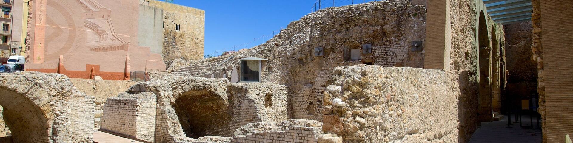 Ruinas antiguas con muros y arcos de piedra, un edificio moderno al fondo, y un cielo azul despejado.