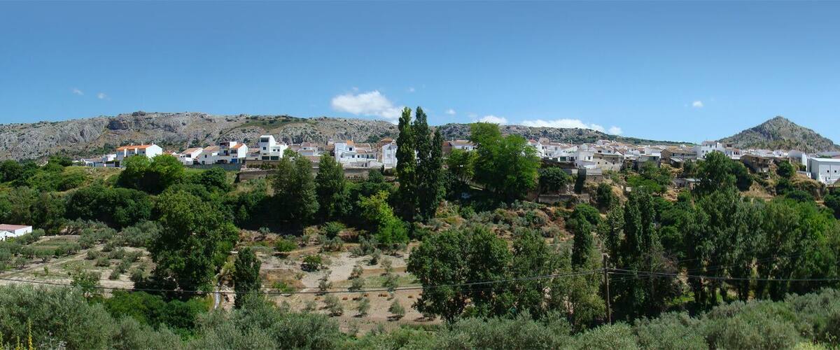 A hillside village with white buildings, dense greenery, and rocky hills in the background.