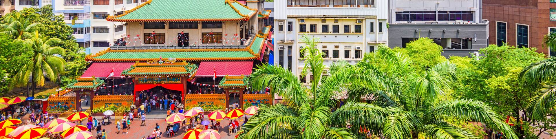 A traditional temple with a green roof, surrounded by palm trees and modern buildings.