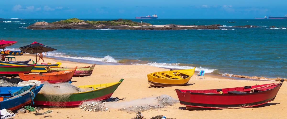 A beach with colorful boats, a rocky outcrop, and a distant ship.