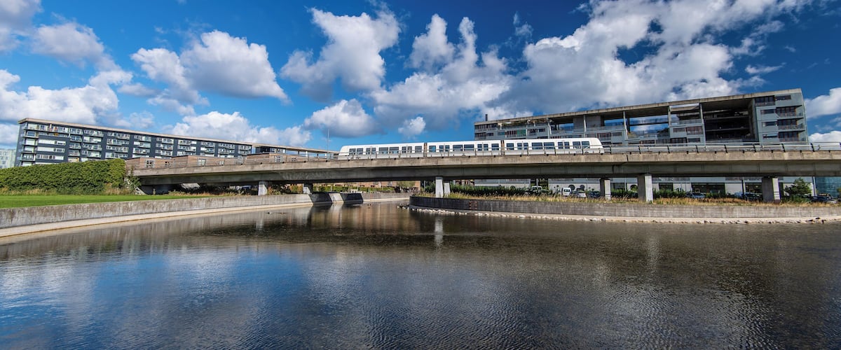 A modern bridge over a calm river with a train crossing, surrounded by greenery and a clear blue sky.